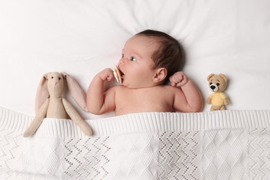 Photo of Cute little baby with toys lying under knitted plaid in bed, top view