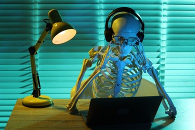 Photo of Waiting concept. Human skeleton sitting at wooden table with laptop and headphones indoors