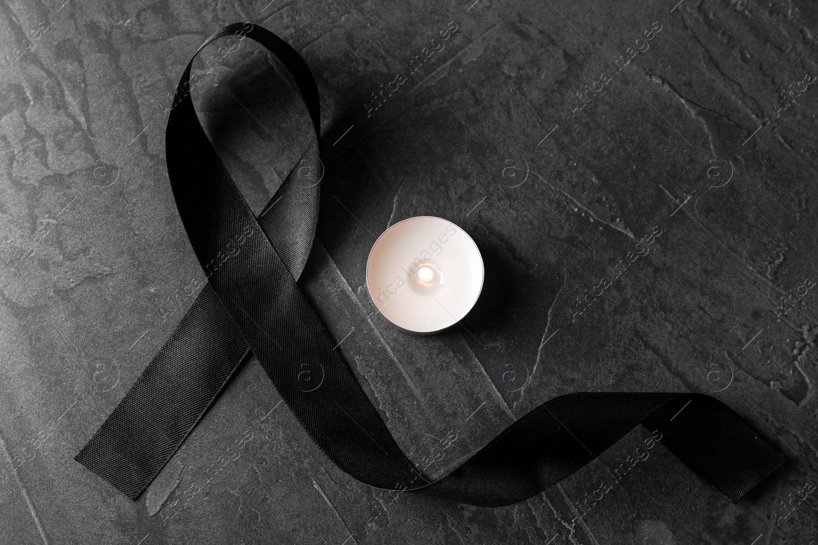 Photo of Black ribbon and burning candle on dark grey stone surface, top view. Funeral symbols