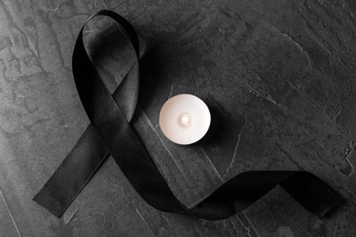 Photo of Black ribbon and burning candle on dark grey stone surface, top view. Funeral symbols