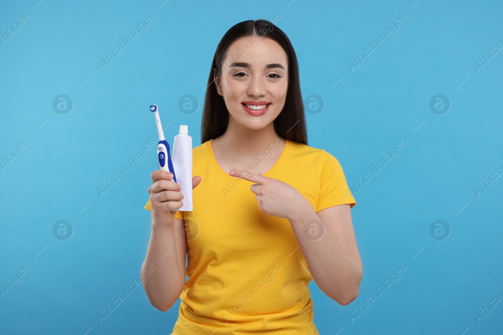 Photo of Happy young woman holding plastic toothbrush and tube of toothpaste on light blue background