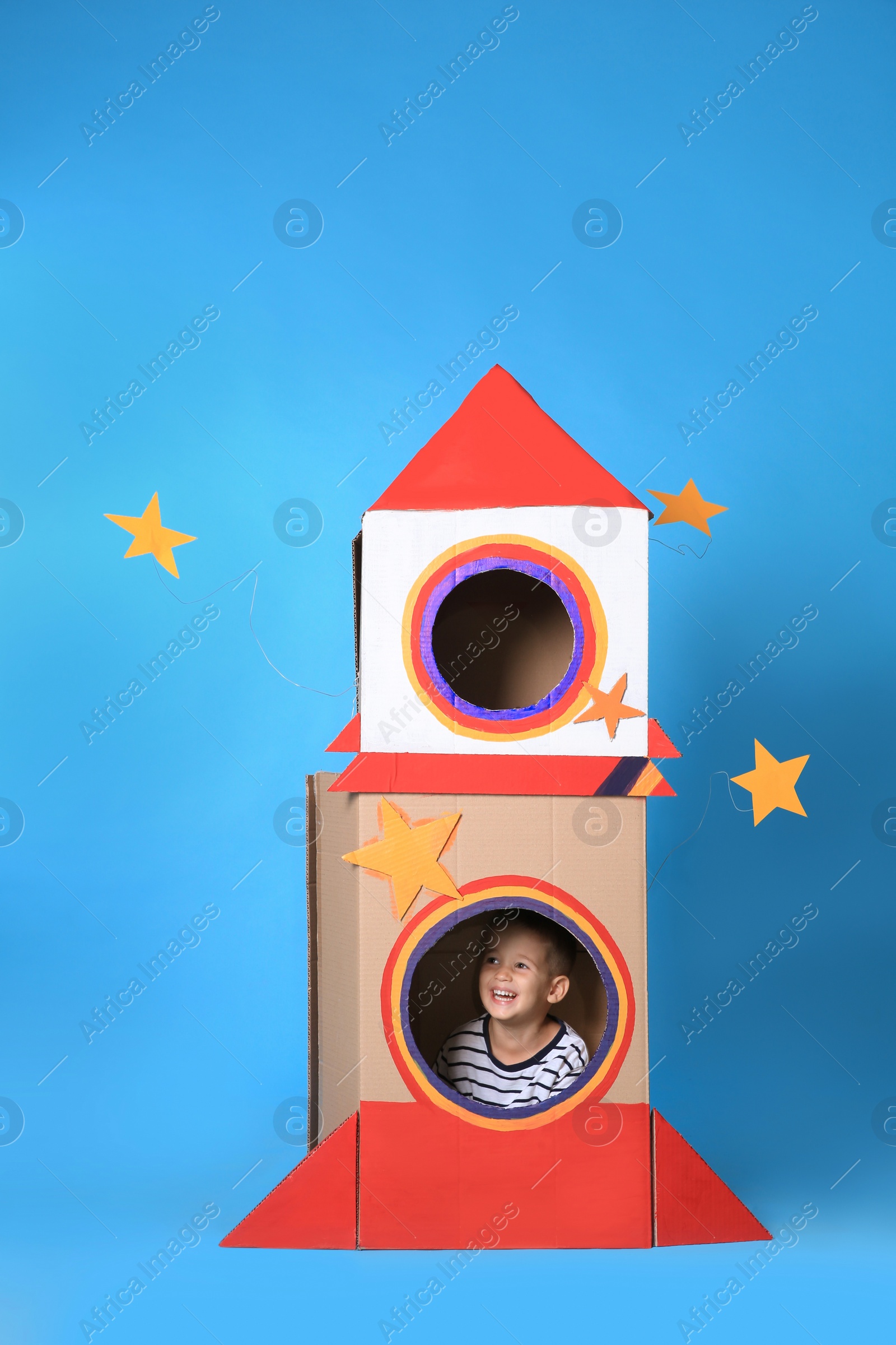 Photo of Cute little boy playing with cardboard rocket on light blue background