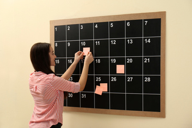 Photo of Young woman putting sticker on board calendar  indoors