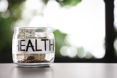 Photo of Glass jar with money and word HEALTH on table against blurred background, space for text
