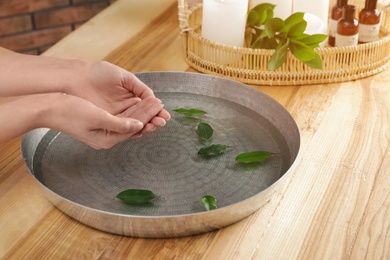 Photo of Woman soaking her hands in bowl with water and leaves on wooden table, closeup. Spa treatment