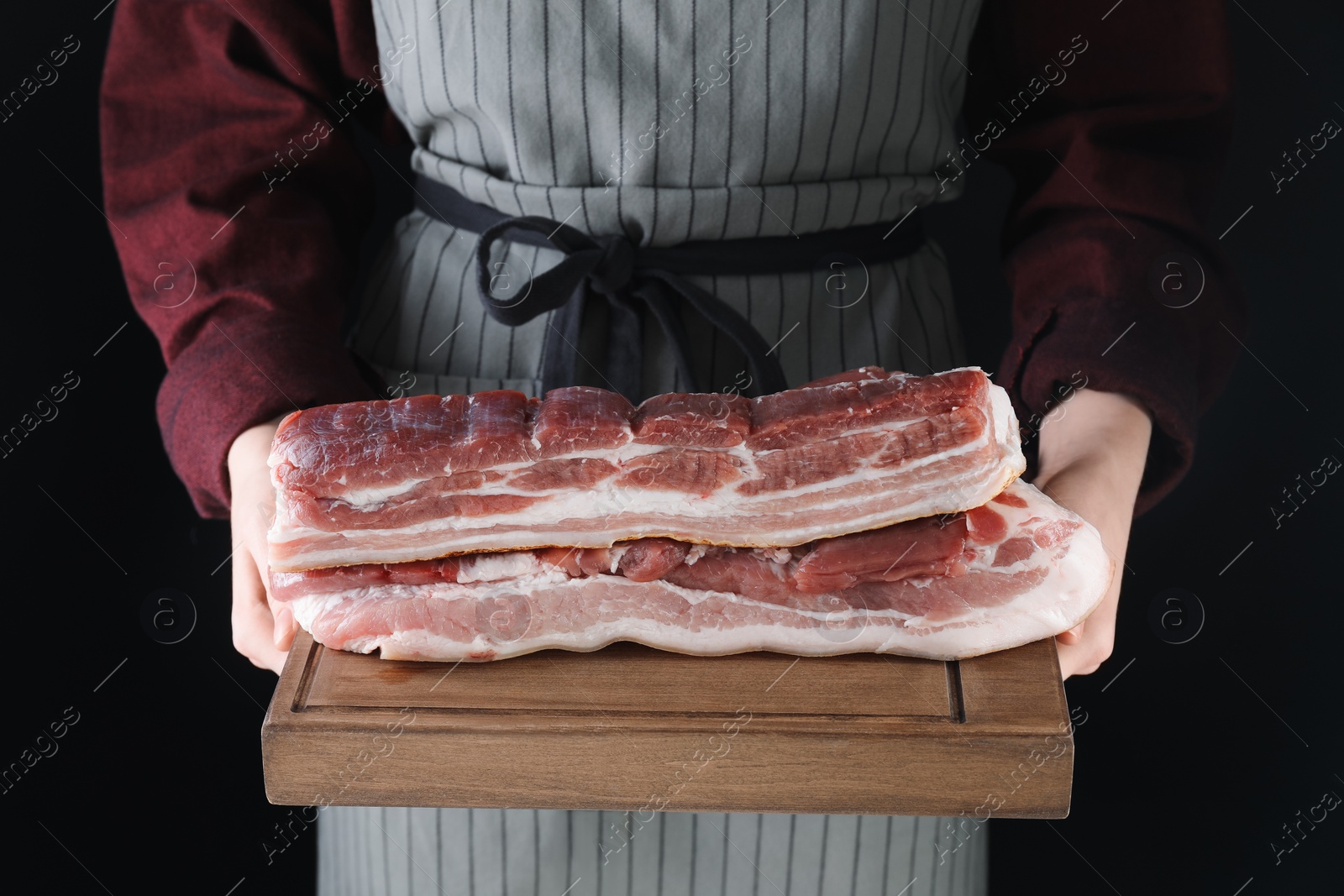 Photo of Woman holding wooden board with pieces of raw pork belly on black background, closeup