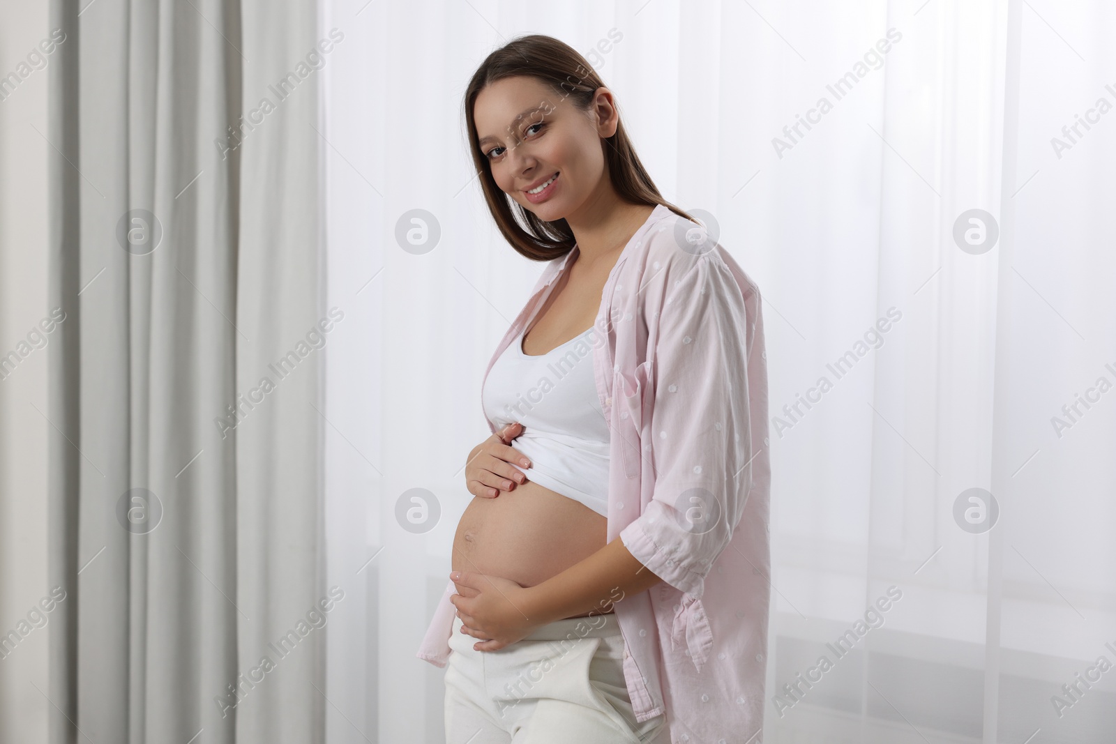 Photo of Beautiful pregnant woman in pink shirt near window indoors