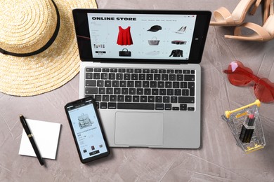 Photo of Online store website on laptop screen. Computer, smartphone, shopping cart, stationery and accessories on grey table, flat lay