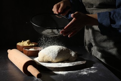 Photo of Woman sprinkling flour over dough at black table, closeup