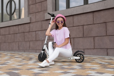 Young woman with cup of coffee sitting on electric kick scooter outdoors