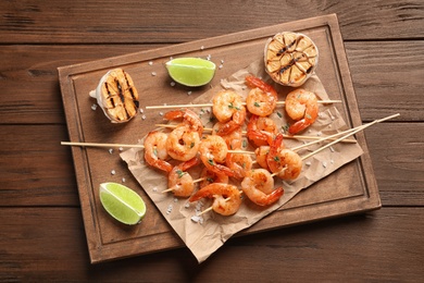 Photo of Board with delicious fried shrimp skewers, lime and garlic on wooden background, top view