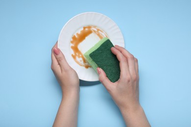 Woman washing dirty plate with sponge on light blue background, top view