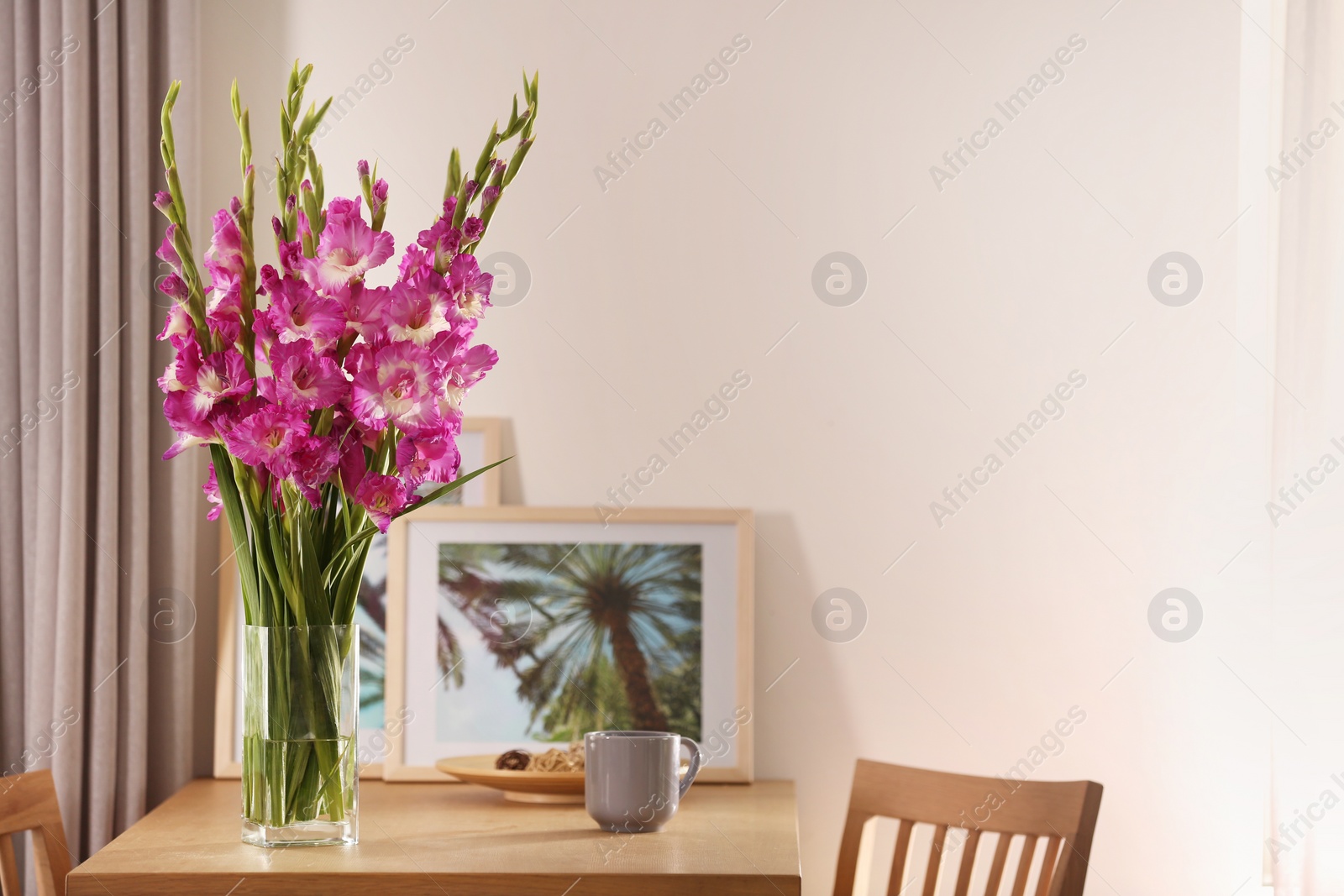 Photo of Vase with beautiful pink gladiolus flowers, pictures and cup on wooden table in room, space for text