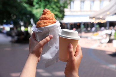 Woman holding croissant and paper cup of coffee on city street, closeup