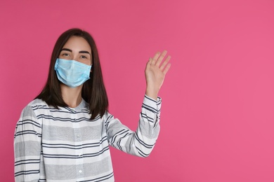 Photo of Young woman in protective mask showing hello gesture on pink background, space for text. Keeping social distance during coronavirus pandemic