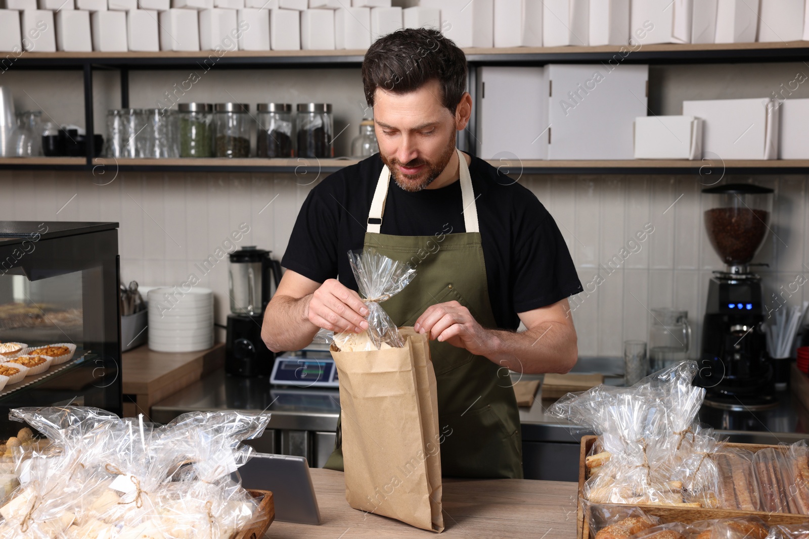 Photo of Smiling seller putting pastry into paper bag at cashier desk in bakery shop