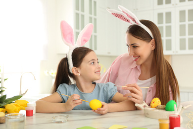 Photo of Happy mother and daughter with bunny ears headbands painting Easter egg in kitchen