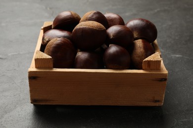 Photo of Roasted edible sweet chestnuts in wooden crate on grey textured table, closeup