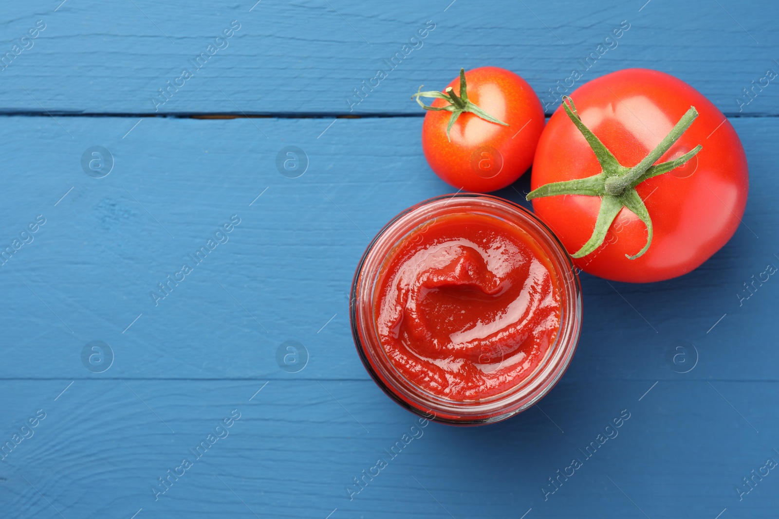 Photo of Jar of tasty ketchup and tomatoes on blue wooden table, flat lay. Space for text