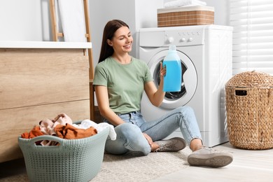Photo of Woman sitting on floor near washing machine and holding fabric softener in bathroom