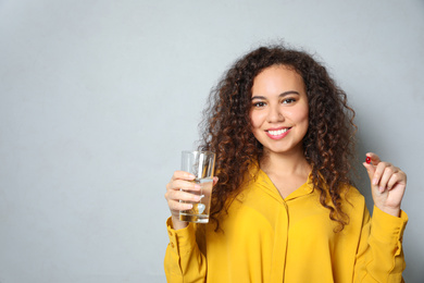 Photo of African-American woman with glass of water and vitamin capsule on light grey background. Space for text