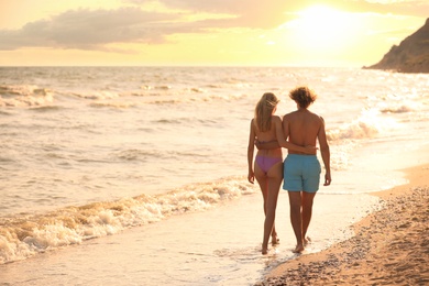 Young woman in bikini and her boyfriend walking on beach at sunset. Lovely couple