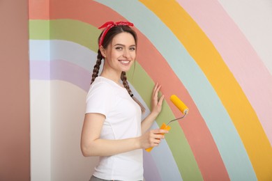 Photo of Young woman holding roller near wall with painted rainbow indoors