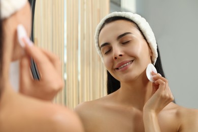 Young woman using cotton pad with micellar water near mirror indoors