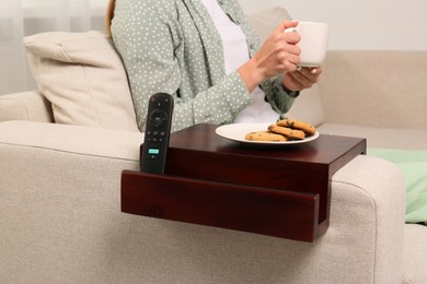 Photo of Remote control and cookies on sofa armrest wooden table. Woman holding cup of drink at home, closeup