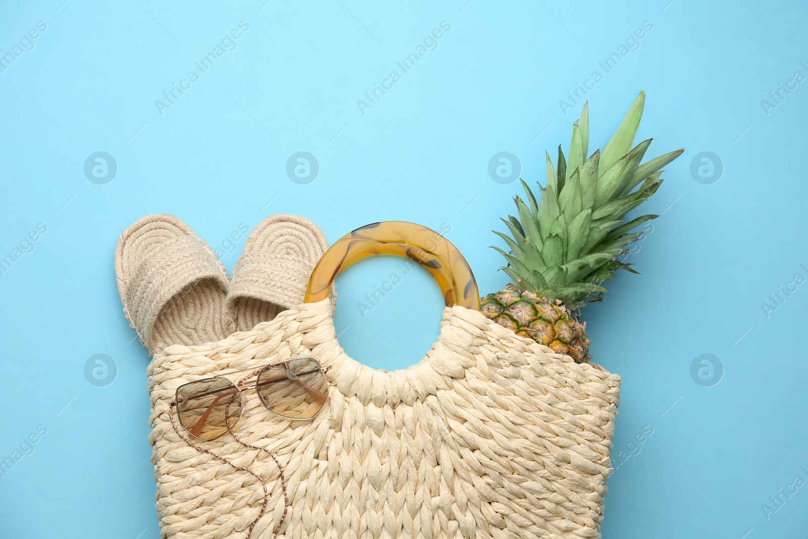 Photo of Elegant woman's straw bag with shoes, sunglasses and pineapple on light blue background, top view