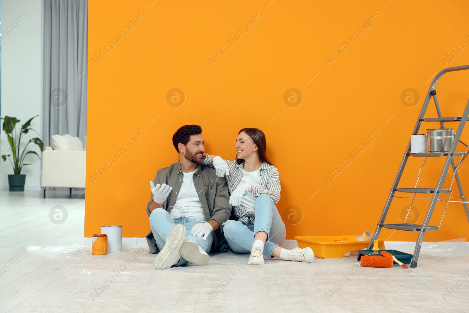 Photo of Happy designers sitting on floor with painting equipment near freshly painted orange wall indoors