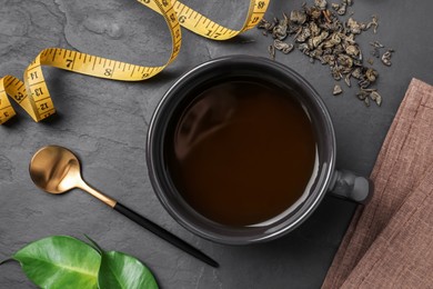 Cup of herbal diet tea and measuring tape on black table, flat lay. Weight loss concept