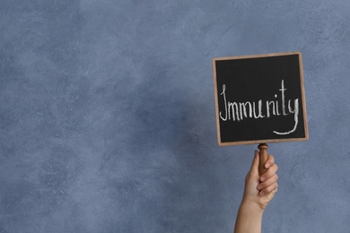 Photo of Woman holding small chalkboard with word Immunity on blue background, closeup. Space for text