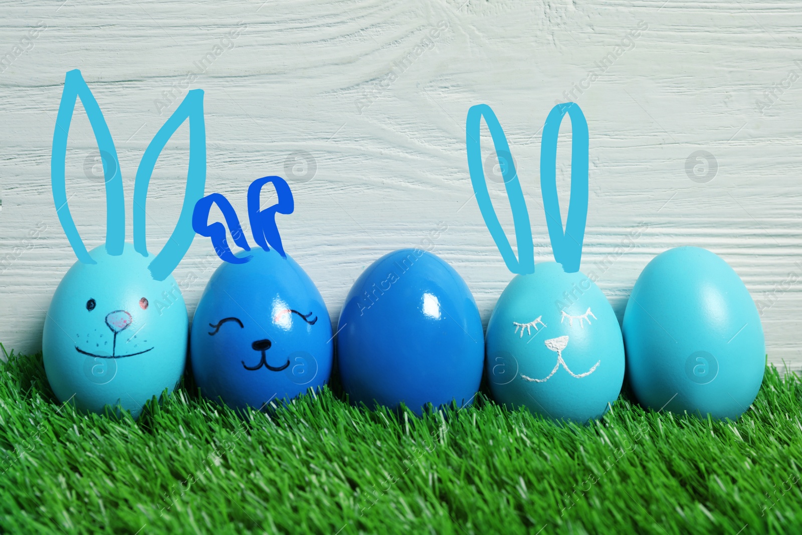 Image of Several eggs with drawn faces and ears as Easter bunnies among others on green grass against white wooden background