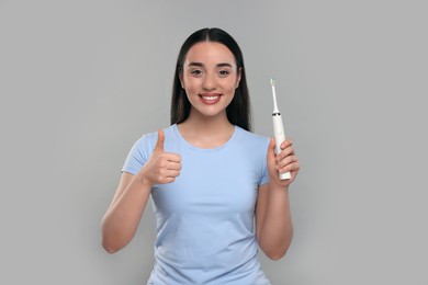 Photo of Happy young woman holding electric toothbrush and showing thumb up on light grey background