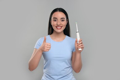 Happy young woman holding electric toothbrush and showing thumb up on light grey background