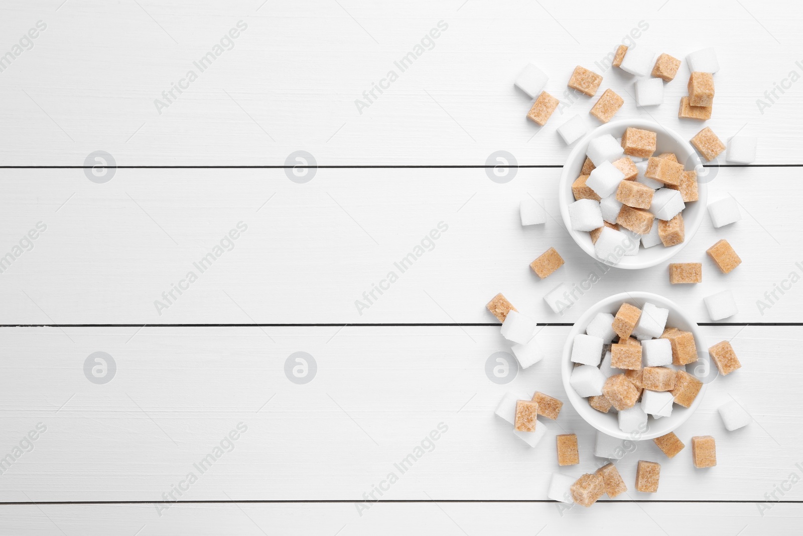 Photo of Different sugar cubes in bowls on white wooden table, flat lay. Space for text