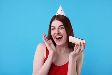 Happy woman in party hat with piece of cake on light blue background