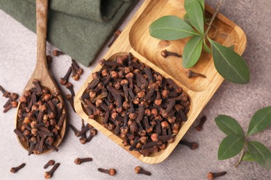 Photo of Wooden tray with aromatic cloves, spoon and green leaves on brown table, flat lay