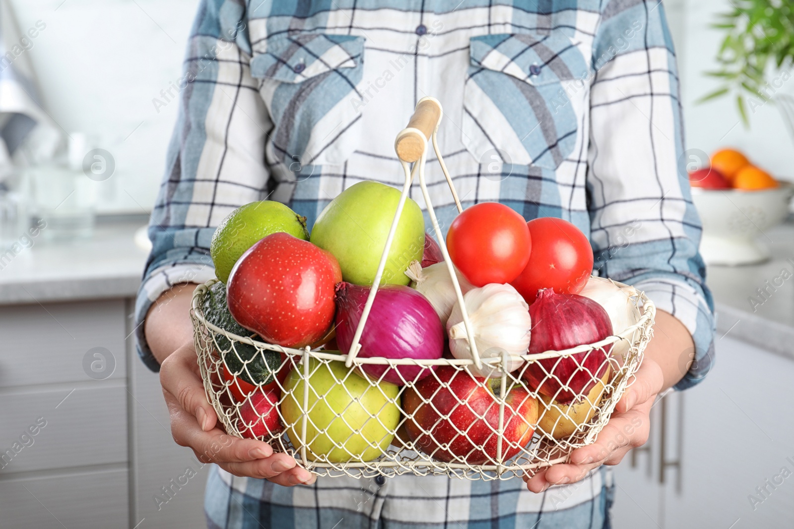 Photo of Woman holding basket full of fresh vegetables and fruits in kitchen, closeup