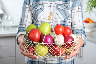 Photo of Woman holding basket full of fresh vegetables and fruits in kitchen, closeup