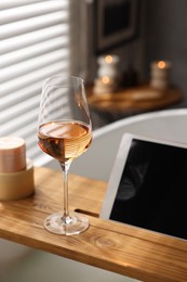 Photo of Wooden tray with tablet and wine on bathtub in bathroom