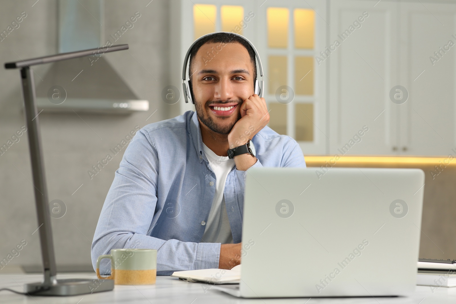 Photo of Young man with headphones working on laptop at desk in kitchen. Home office