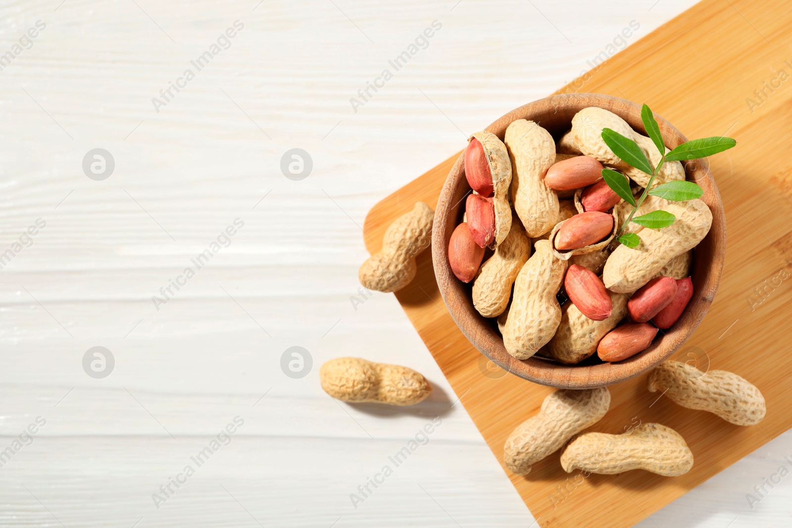 Photo of Fresh unpeeled peanuts in bowl and twig on white wooden table, top view. Space for text