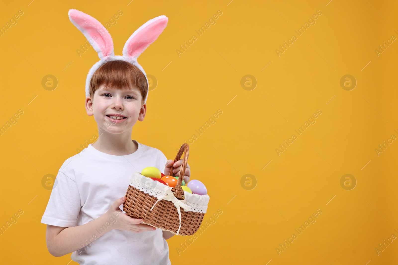 Photo of Easter celebration. Cute little boy with bunny ears and wicker basket full of painted eggs on orange background. Space for text