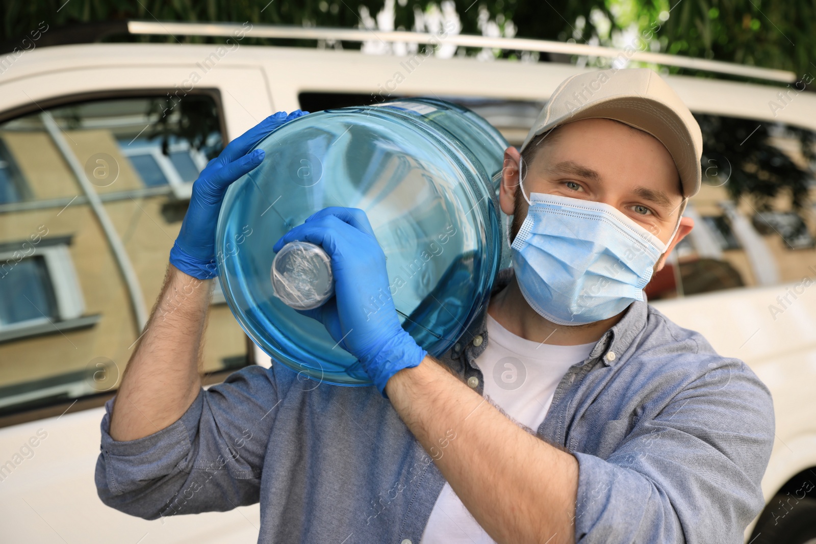 Photo of Courier in medical mask holding bottle of cooler water near car outdoors. Delivery during coronavirus quarantine