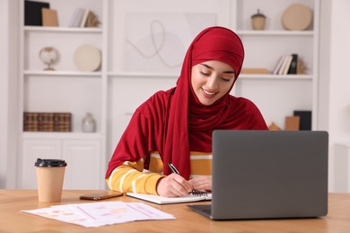 Muslim woman writing notes near laptop at wooden table at home