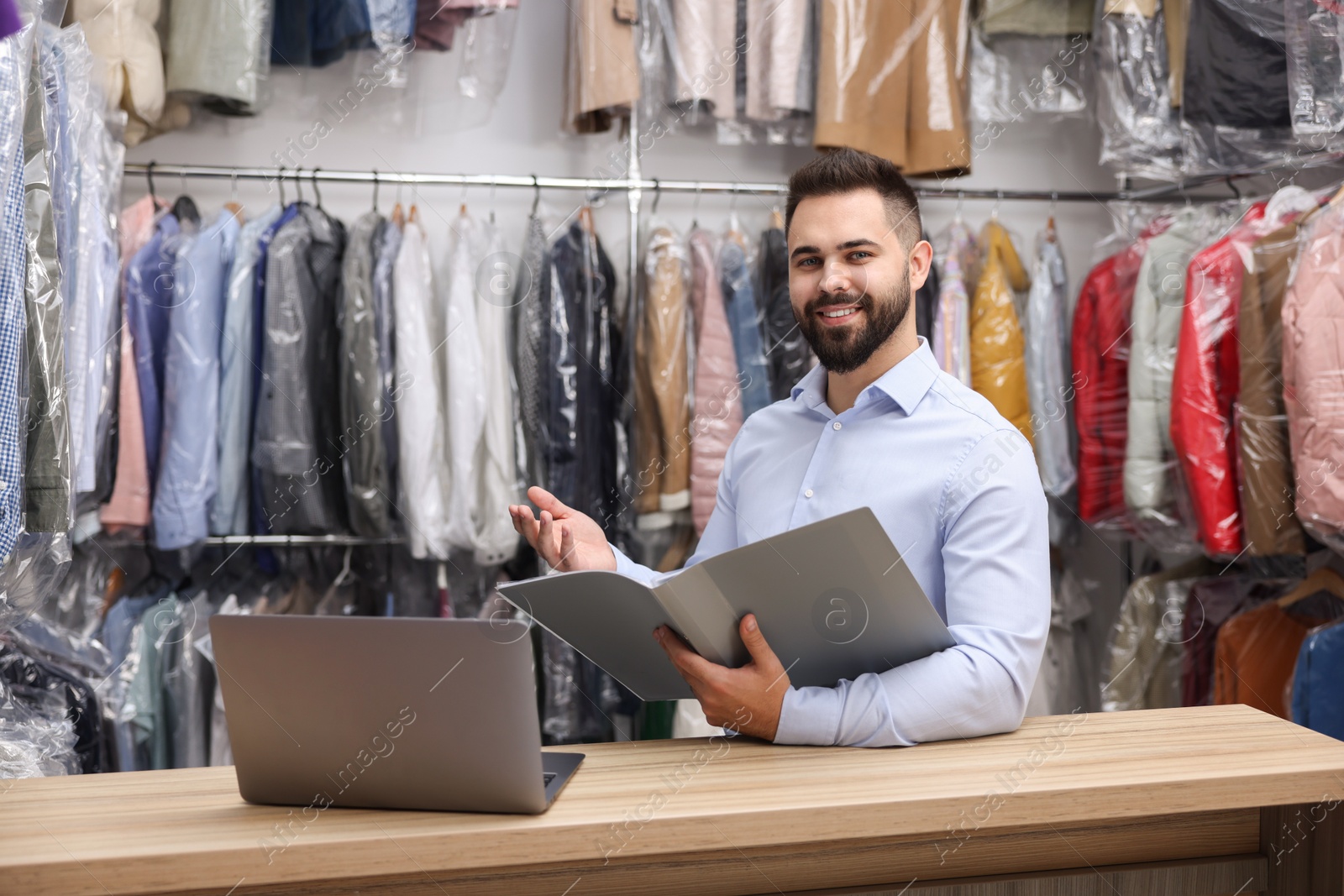 Photo of Dry-cleaning service. Happy worker with folder and laptop at counter indoors