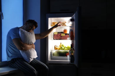 Photo of Sleepy man with pillow near refrigerator in kitchen at night