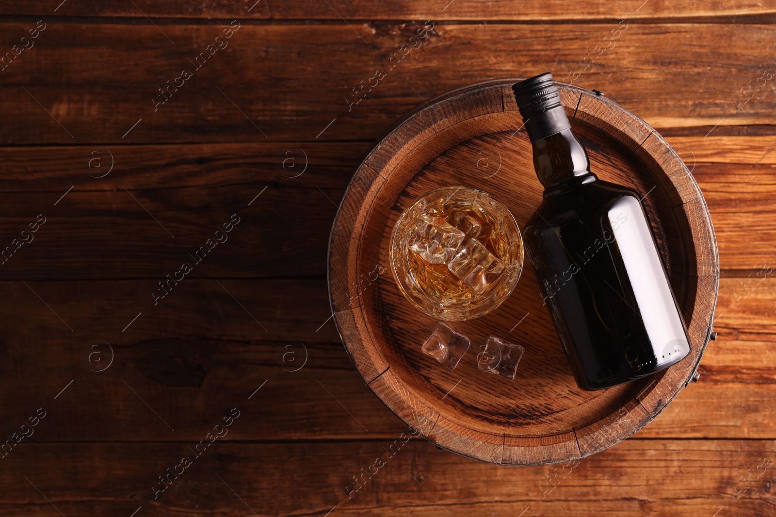 Photo of Whiskey with ice cubes in glass, bottle and barrel on wooden table, top view. Space for text